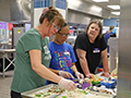 Food Service staff members pit vegetables on mini pizzas during a cooking lesson.